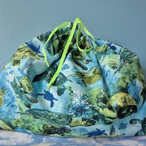 May include: A drawstring bag with a sea turtle pattern. The bag features a blue background with green and yellow sea turtles. The drawstring is a bright neon green. The bag is made of fabric.