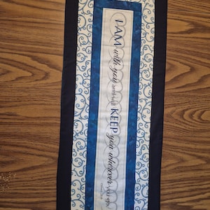 May include: A rectangular fabric wall hanging featuring a central white panel with the text "I AM with you and will KEEP you wherever you go. Genesis 28:15" in black, bordered by blue mottled fabric and a white scrollwork pattern on blue.
