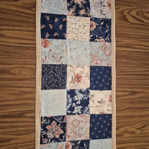 May include: A rectangular patchwork quilt featuring a variety of floral and paisley patterns in shades of blue, cream, and peach. The quilt is bordered with a solid tan fabric. The design is a mix of squares and rectangles, creating a traditional, handcrafted look.