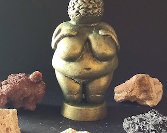 May include: A gold-colored figurine of a woman with a round head and a large, round body. The figurine is standing on a small, round base. The figurine is surrounded by small, brown, gray, and white rocks.
