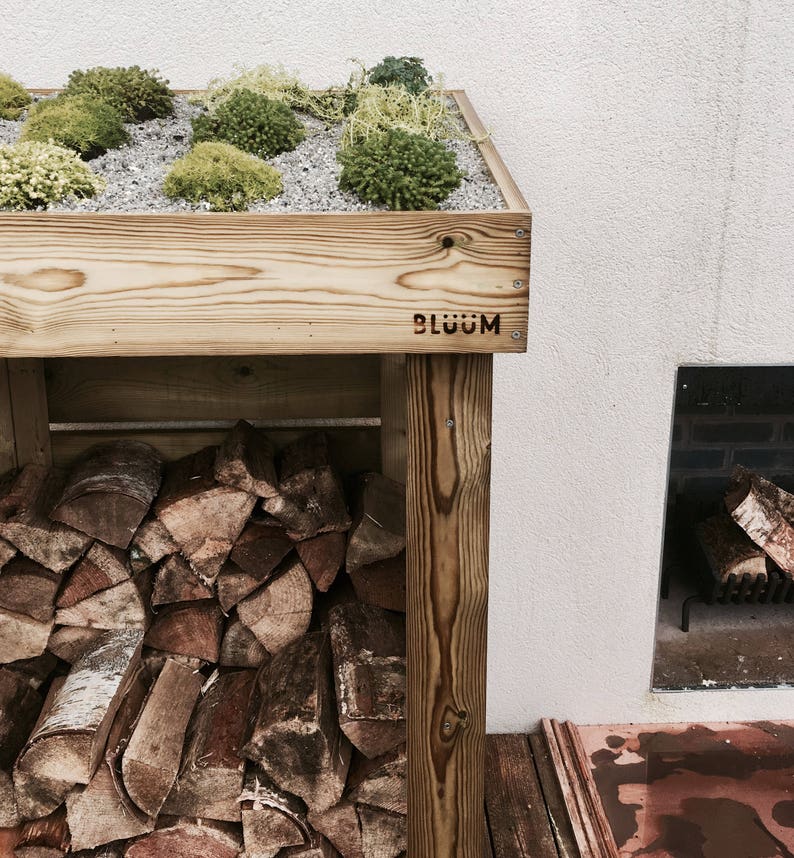 May include: A wooden log rack with a green roof. The roof is covered in small plants and gravel. The rack is labeled "BL&Uuml;&Uuml;M".