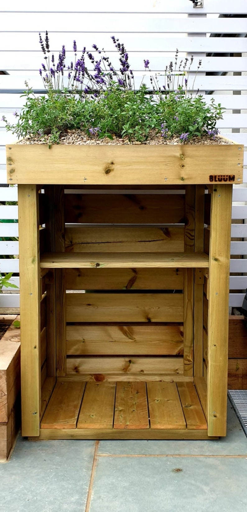 May include: A wooden shelf unit with two shelves and a planter box on top. The planter box is filled with lavender and other plants. The shelf unit is labeled "BLOOM".