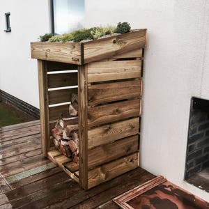 May include: A wooden log storage rack with a green roof. The rack has a slanted roof and is made of natural wood. The rack is filled with logs and is placed against a white wall.