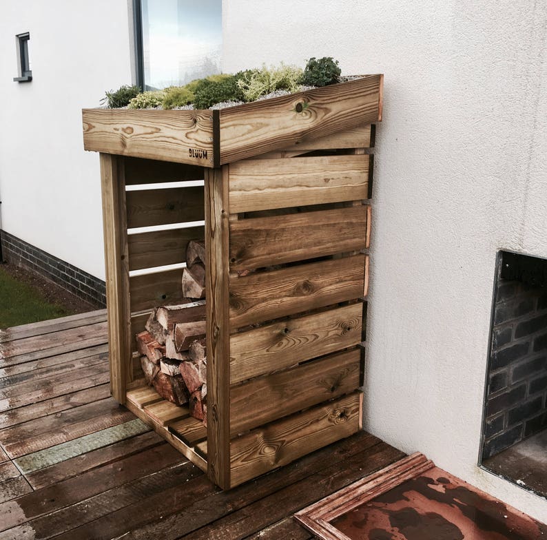 May include: A wooden log storage rack with a green roof. The rack has a slanted roof and is made of natural wood. The rack is filled with logs and is placed against a white wall.