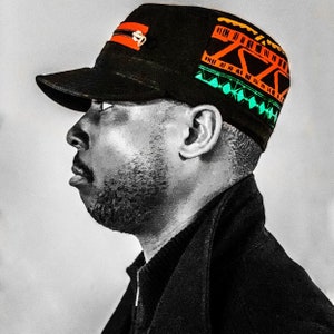 May include: A black and green patterned baseball cap with a zipper detail and orange accents. The cap is worn by a person with a beard.