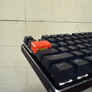 May include: A black mechanical keyboard with a red miniature container on the Caps Lock key. The keyboard has black keycaps and a silver frame. The container is rectangular and has a red exterior.