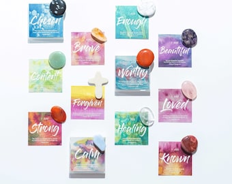 May include: A set of 12 affirmation cards with watercolour backgrounds and inspirational messages. Each card features a different affirmation, such as "I am strong", "I am loved", and "I am enough". Each card has a corresponding stone or crystal on top.
