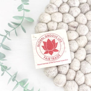 Op de afbeelding: Een wit vilt bal tapijt met een label met de tekst "Global Groove Life Fair Trade" met een rood lotusbloemlogo. Het label vermeldt ook "Proud member of the Fair Trade Federation."