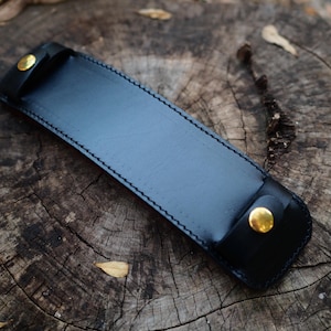 May include: A black leather sheath with gold-coloured button closures. The rectangular case has a stitched border and is designed to hold a long, thin object. The sheath is displayed on a weathered wooden surface.