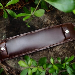 May include: A dark brown leather item, possibly a sheath or holder, with decorative stitching along the edges. The item has a rectangular shape with rounded ends and is secured with silver-coloured rivets. The background is a wooden surface with green foliage.