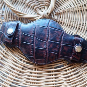 May include: A dark brown leather holster with a crocodile skin effect. The holster has a curved shape with a press stud closure on each end. The background is a woven basket.