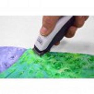May include: A white and black electric trimmer is used to cut a piece of green and blue fabric. The fabric has a watercolor effect. The trimmer is held by a person's hand.