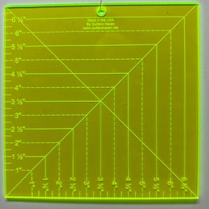 May include: A neon yellow quilting ruler with black markings and measurements in inches. The ruler is square, with diagonal and horizontal lines for precise cutting. Text on the ruler reads "Made in the USA By Quilters Haven www.quiltershaven.net".