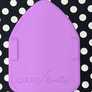 May include: A lavender-colored, heart-shaped sewing mat with two side tabs. The mat features the text "QUILTED heartz" with a dotted line and heart design. The mat is on a black and white polka dot background.