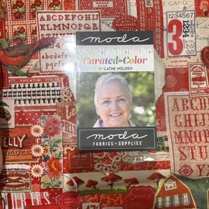 May include: A fabric swatch featuring a collage of vintage-style red and orange graphics, including text like SOAP, alphabet letters, numbers, and illustrations of birds, mushrooms, and a house, with a Moda Curated in Colour label by Cathe Holden.