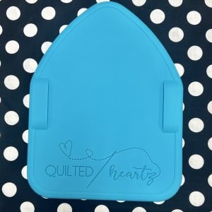 May include: A turquoise-colored, house-shaped sewing template with the text "QUILTED heartz" and a heart design. The template has two side tabs and is set against a black and white polka dot background.