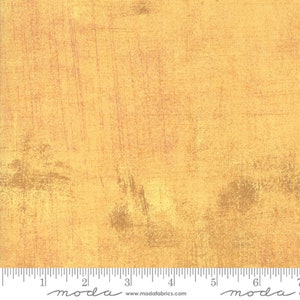 May include: A close-up of a yellow fabric with a textured, distressed look. The fabric has a subtle, faded pink and brown pattern throughout. The fabric is measured in inches with a ruler at the bottom of the image.