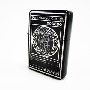 May include: Black lighter with a silver engraved image of Dark Magician Girl from the Yu-Gi-Oh! Trading Card Game. The card text reads "SPELLCASTER/EFFECT" and "ATK/2000 DEF/1700".