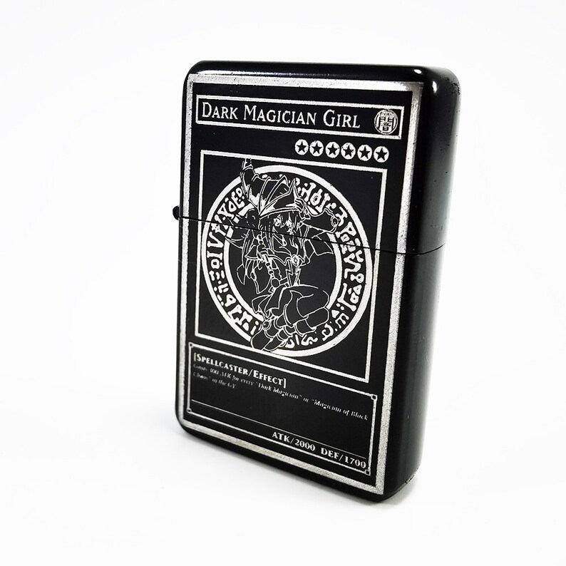 May include: Black lighter with a silver engraved image of Dark Magician Girl from the Yu-Gi-Oh! Trading Card Game. The card text reads "SPELLCASTER/EFFECT" and "ATK/2000 DEF/1700".