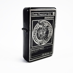 May include: Black lighter with a silver engraving of a Dark Magician Girl Yu-Gi-Oh! card. The card features a black and white illustration of the character with the text "DARK MAGICIAN GIRL" and "SPELLCASTER/EFFECT".
