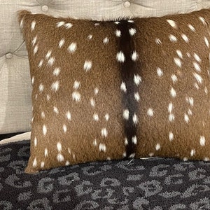May include: A brown and white spotted pillow with a black stripe down the center. The pillow is made of real animal fur and is sitting on a black and gray patterned surface.