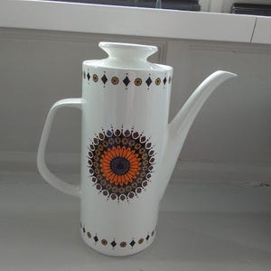 Peut inclure: Pichet à café en céramique blanche avec un long bec et une anse. Le pot présente un grand motif floral orange et marron au centre, avec des motifs décoratifs orange, marron et noir en haut et en bas. Le pot est sur une surface blanche.