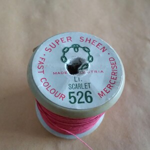 Könnte beinhalten: Eine Spule pinkfarbenen Fadens mit der Aufschrift "Super Sheen Fast Colour Mercerised Made Austria Li. Scarlet 526"