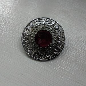 Peut inclure: Épingle de kilt argentée avec un bijou violet foncé au centre et un motif de chardon sur le bord.