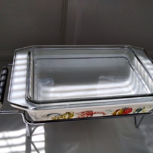May include: A white glass baking dish with a colourful seafood design sits on a silver metal stand. The dish has a clear glass lid.