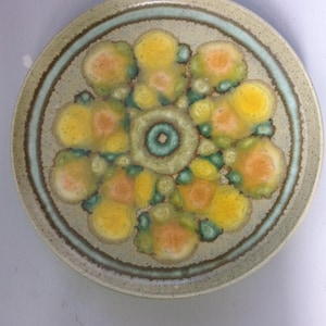 May include: A ceramic plate featuring a floral design in shades of yellow, orange, green, and blue. The plate has a speckled glaze and a raised rim.