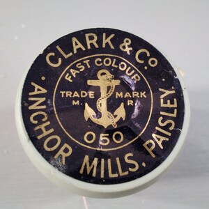 Könnte beinhalten: Eine runde, cremefarbene Garnspule mit einem marineblauen Etikett. Das Etikett zeigt goldenen Text und ein Ankerlogo mit der Aufschrift "CLARK & CO FAST COLOUR ANCHOR MILLS PAISLEY". Es zeigt auch "TRADE MARK" und "050". Wahrscheinlich eine Garnspule.