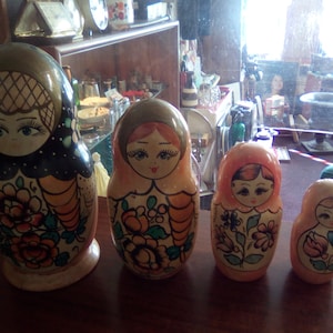 Puede incluir: Cuatro muñecas rusas de madera, también conocidas como muñecas matrioska, se exhiben sobre una superficie de madera oscura. Las muñecas están pintadas con diseños florales y presentan rostros femeninos. La muñeca más grande es azul oscuro, las otras son naranjas y melocotón.