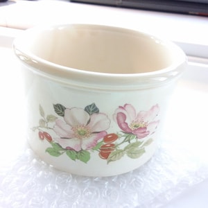 May include: Cream ceramic bowl decorated with a floral pattern. The design includes pink flowers, green leaves, and red berries. The rim and interior are a lighter cream shade. The bowl is on bubble wrap.