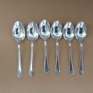 May include: Six polished silver spoons. Each spoon has a long, slender handle and a rounded bowl. The handles have a decorative design near the base and are engraved with the letter "F".