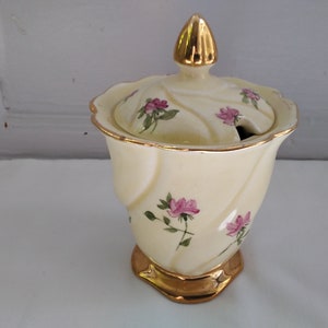 May include: A white ceramic sugar bowl with a lid and gold trim. The bowl is decorated with pink roses and green leaves. The lid has a gold knob.