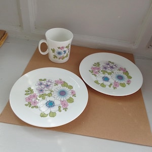 Könnte beinhalten: Ein weißer Keramikbecher und zwei Teller mit einem Blumendesign. Die Blumen sind weiß, blau und rosa mit grünen Blättern. Der Becher hat einen Henkel und die Teller sind rund.