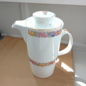 May include: White ceramic coffee pot with a lid and handle. The pot is decorated with a floral pattern in pink, yellow, and blue. The rim and handle are trimmed with gold. The pot is approximately 8 inches tall.