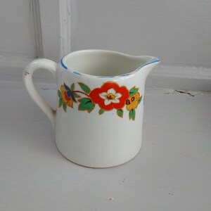 Könnte beinhalten: Weißer Keramik-Sahnegießer mit blauem Rand und Griff. Der Sahnegießer ist mit einem Band aus bunten Blumen in Rot, Gelb und Blau verziert. Der Sahnegießer ist wahrscheinlich Vintage.