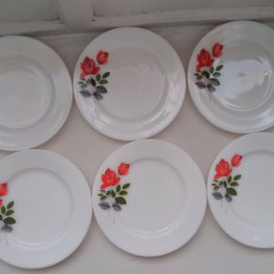 May include: Six white ceramic plates with a red rose design. The plates have a slightly raised rim and a smooth surface.