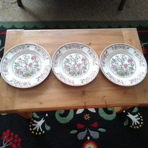 May include: Three white ceramic plates with a floral design. The plates feature a detailed pattern of pink, blue, and green flowers and foliage. The plates are arranged on a wooden table.