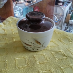 May include: A vintage ceramic sugar bowl with a brown lid and a cream-colored base. The base features a wheat stalk design. The bowl is sitting on a yellow textured cloth.