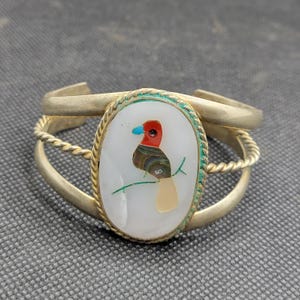 May include: A metal cuff bracelet featuring a central oval inlay of white shell. The inlay displays a bird motif crafted from inlaid pieces of red, brown, and tan stone, with a small blue beak. The bracelet has a twisted rope border and band.