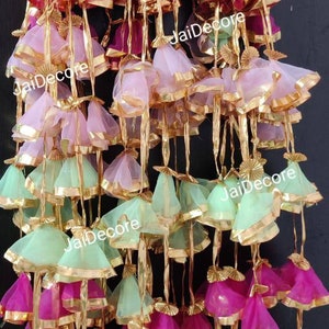 May include: A collection of decorative hanging tassels made of pink, green, and white sheer fabric with gold trim. The tassels are arranged in rows and are hanging from a white bar.