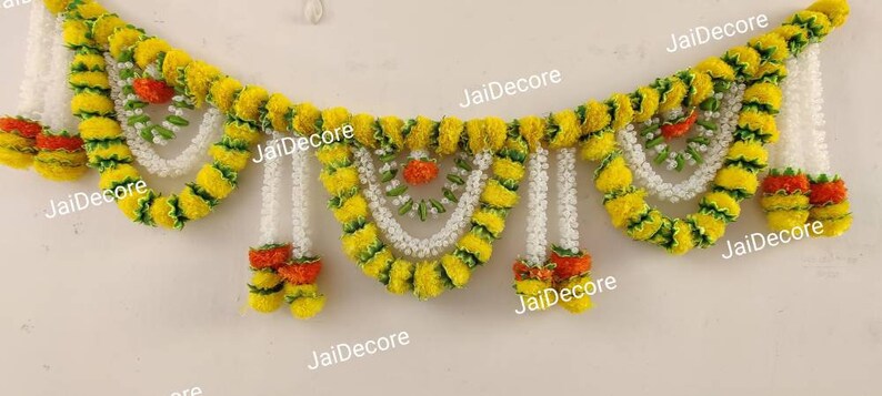 May include: A yellow and green garland made of artificial flowers with white beads hanging from it. The garland is decorated with orange accents and has the text "JaiDecor" printed on it.