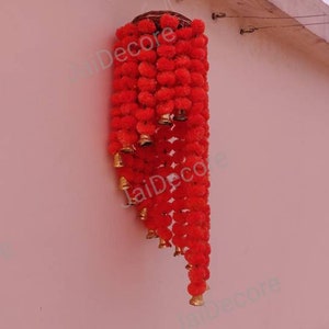 May include: A red flower garland with gold bells hanging from a wall. The garland is made of artificial flowers and is a popular decoration for Indian weddings and festivals.