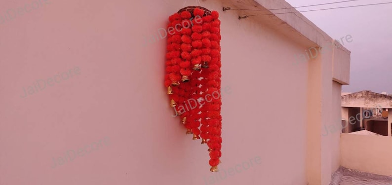 May include: A red flower garland with gold bells hanging from a wall. The garland is made of artificial flowers and is a popular decoration for Indian weddings and festivals.