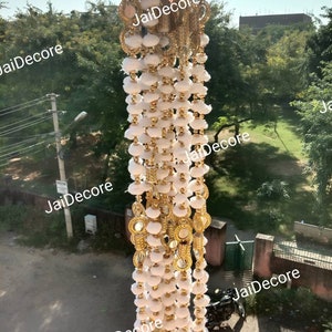 Puede incluir: Pompones blancos con ribetes dorados y pequeños espejos redondos cuelgan de una cuerda. La cuerda está colgada de una ventana y el fondo es una vista borrosa de árboles y edificios.