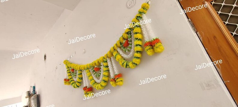 May include: A yellow and white floral garland with green accents hangs on a white wall. The garland is made of artificial flowers and is decorated with the text "JaiDecor" repeated multiple times.