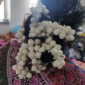 Pu&ograve; includere: Un primo piano di un accessorio per capelli fatto di fiori di stoffa bianca. I fiori sono disposti a grappolo e sono attaccati a un elastico per capelli nero. L'accessorio per capelli &egrave; indossato da una persona con i capelli neri.
