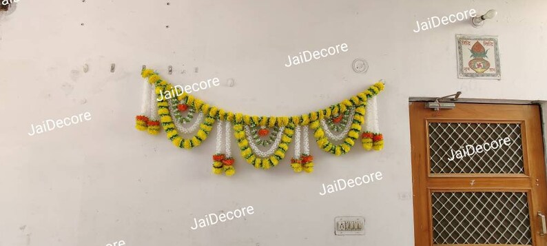 May include: A yellow and white floral garland with green accents hangs on a white wall. The garland is made of artificial flowers and is decorated with small, orange flowers. The garland is a traditional Indian decoration used for festivals and celebrations.